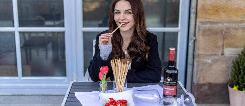 Frau sitzt an einem Tisch auf einer Terrasse mit Wein, Gebäckstangen und kleinen Snacks.