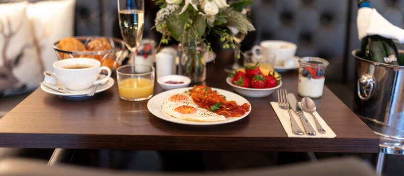 Gedeckter Frühstückstisch mit Spiegeleiern, Speck, Brötchen, Erdbeeren, Kaffee, Orangensaft und einem Glas Sekt.