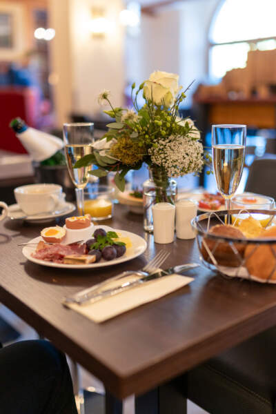 Gedeckter Frühstückstisch mit Brötchen, Ei, Käse, Aufschnitt, Obst, Kaffee, Sekt und frischem Blumenstrauß.