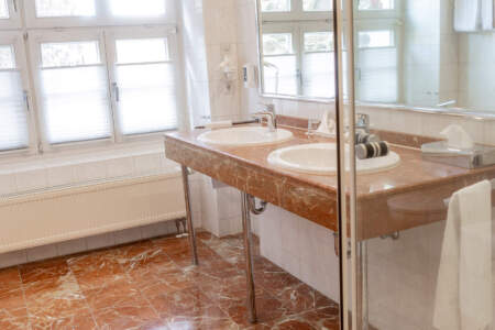 Helles Hotelbad mit zwei Waschbecken, großem Spiegel, Dusche und rötlichem Natursteinboden