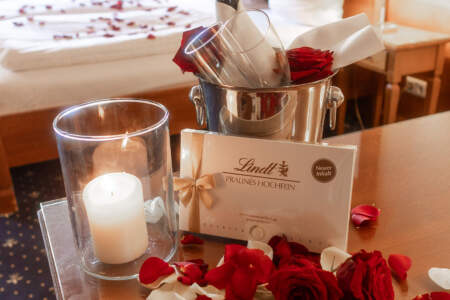 Romantisches Arrangement im Hotelzimmer mit Rosenblättern, roten Rosen, Pralinen, Kerze und Sektkühler vor dem Bett.
